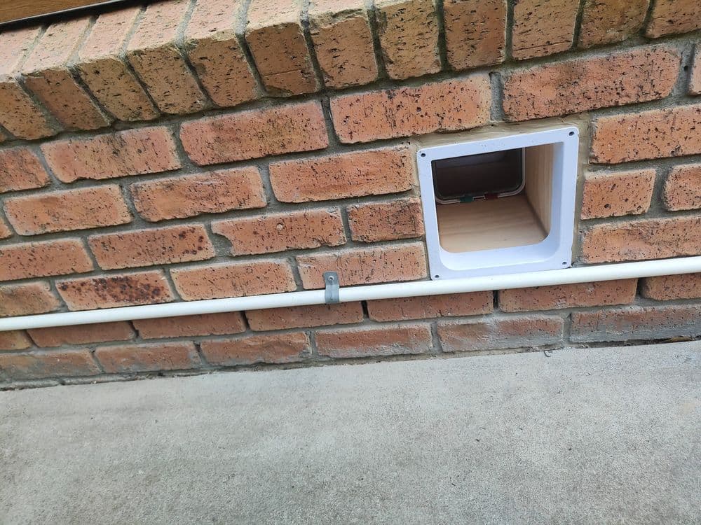 A white cat door installed through a brick wall into an enclosure