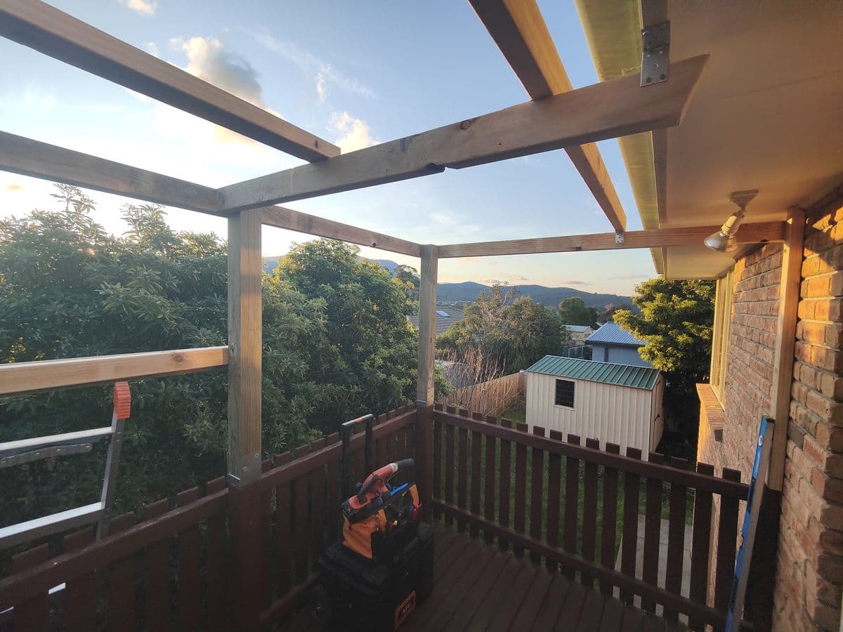 A new timber frame rising from a deck beside a brick house, with a tool bag on the deck and trees beyond
