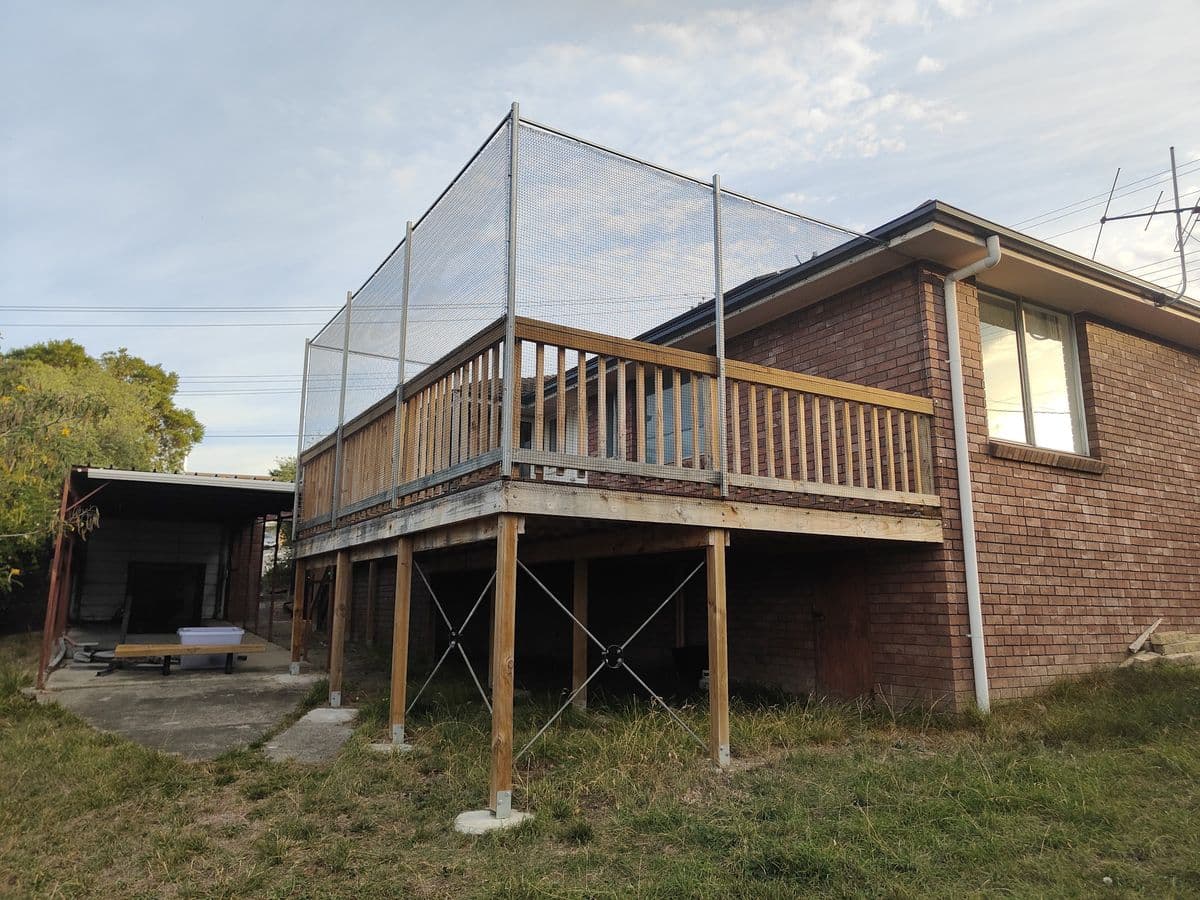 A raised timber deck on the back of a brick house, enclosed with tall netting panels on posts