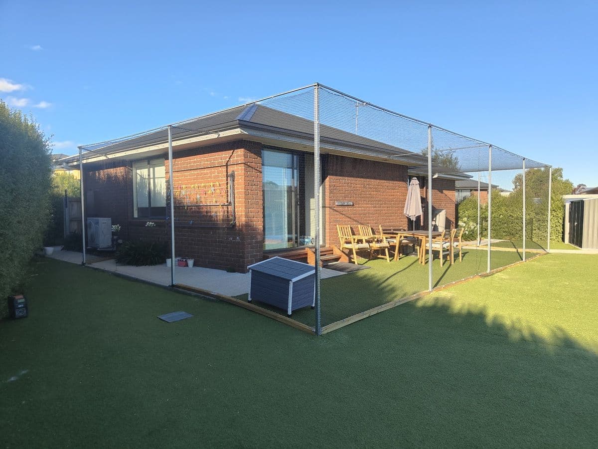 Netting on tall posts enclosing the turfed back yard and outdoor dining area of a brick house under a clear blue sky