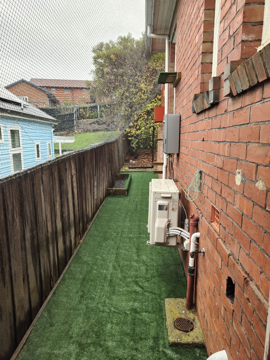 A narrow netted run floored with artificial turf between a red brick house and a timber paling fence