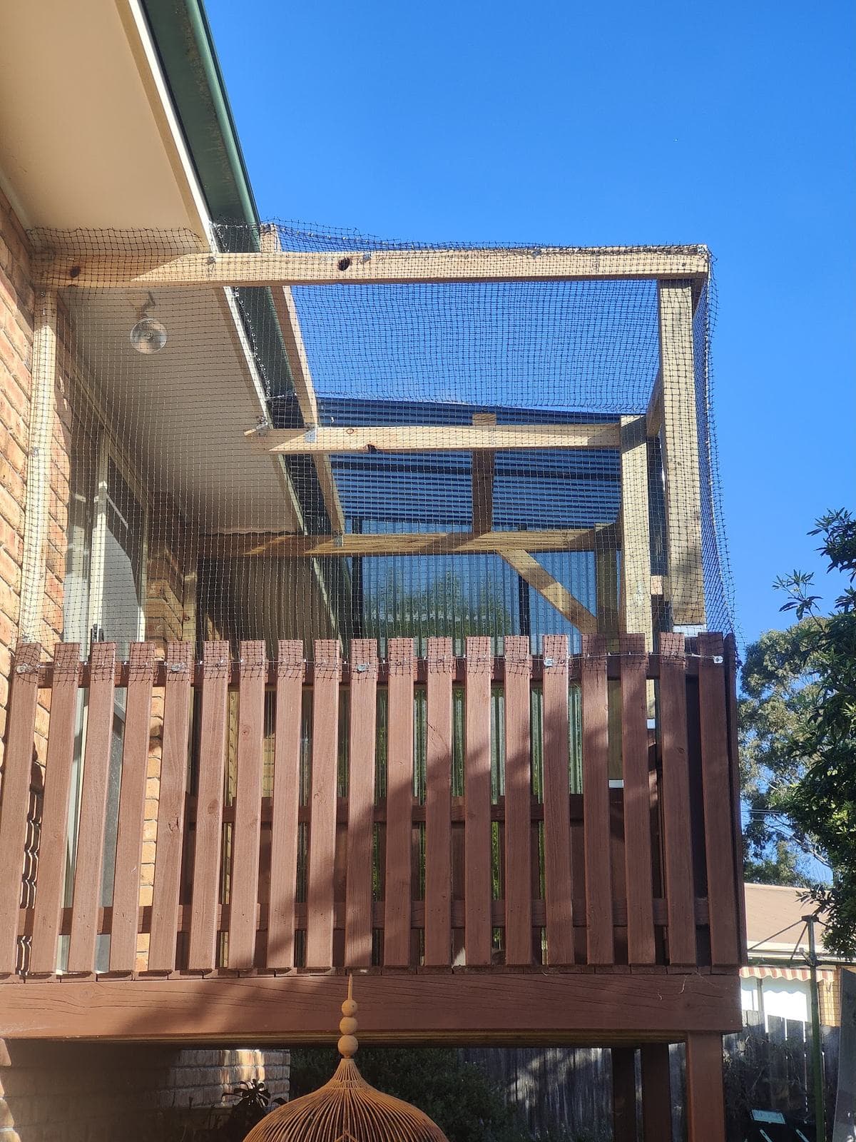 A raised timber balcony on a brick house, enclosed with a timber frame and fine netting under a clear blue sky