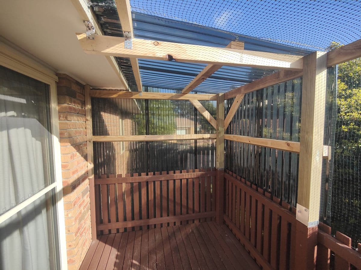 A timber-framed catio on a deck beside a sliding door, with mesh walls, timber slat screens and a clear corrugated roof