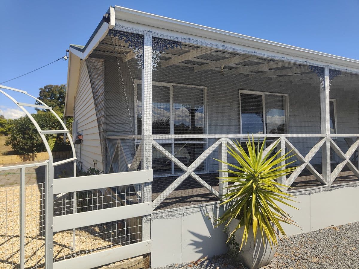 The front verandah of a grey weatherboard house enclosed in fine white mesh behind white cross-braced railings
