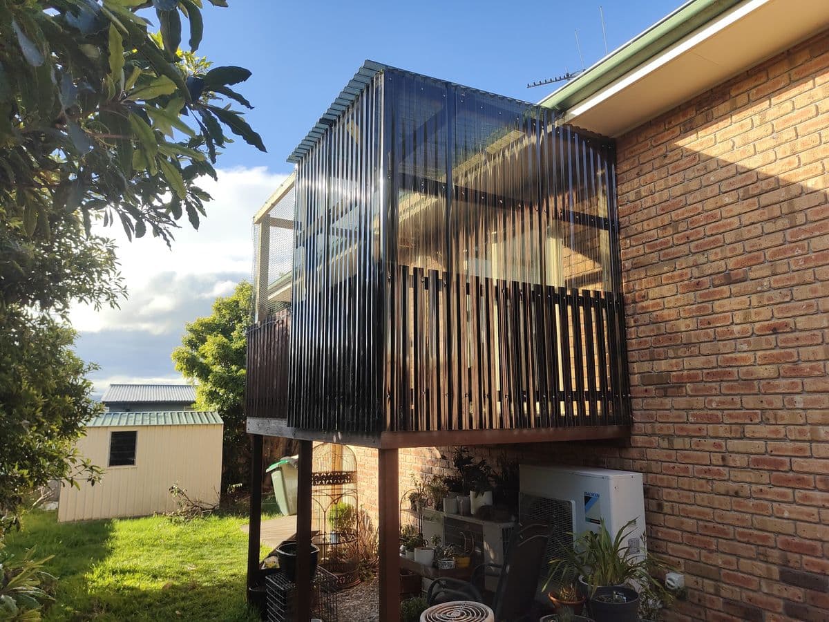 A raised timber deck catio on a brick house with dark slat screens, a roof and mesh walls above the garden