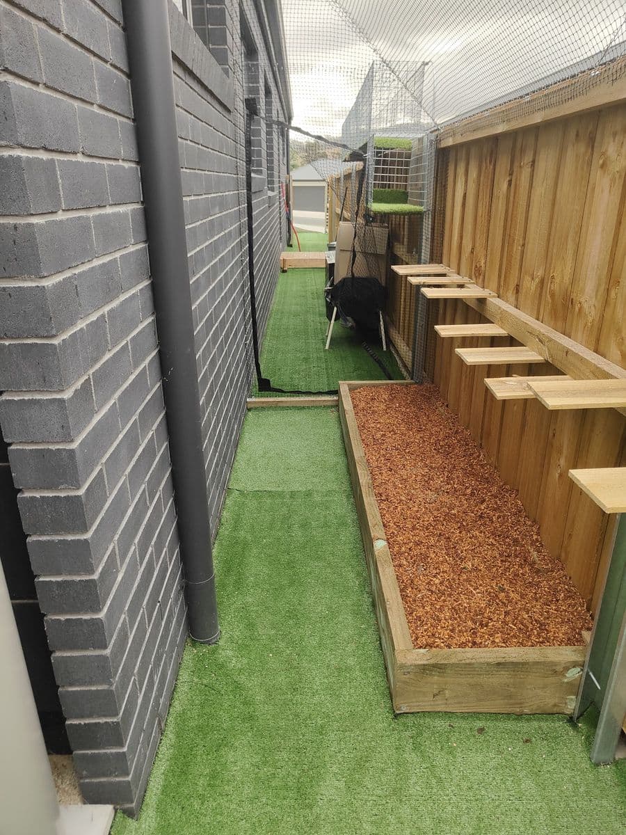 A side run floored with artificial turf beside a dark brick house, with a mulched garden bed and timber steps up the fence