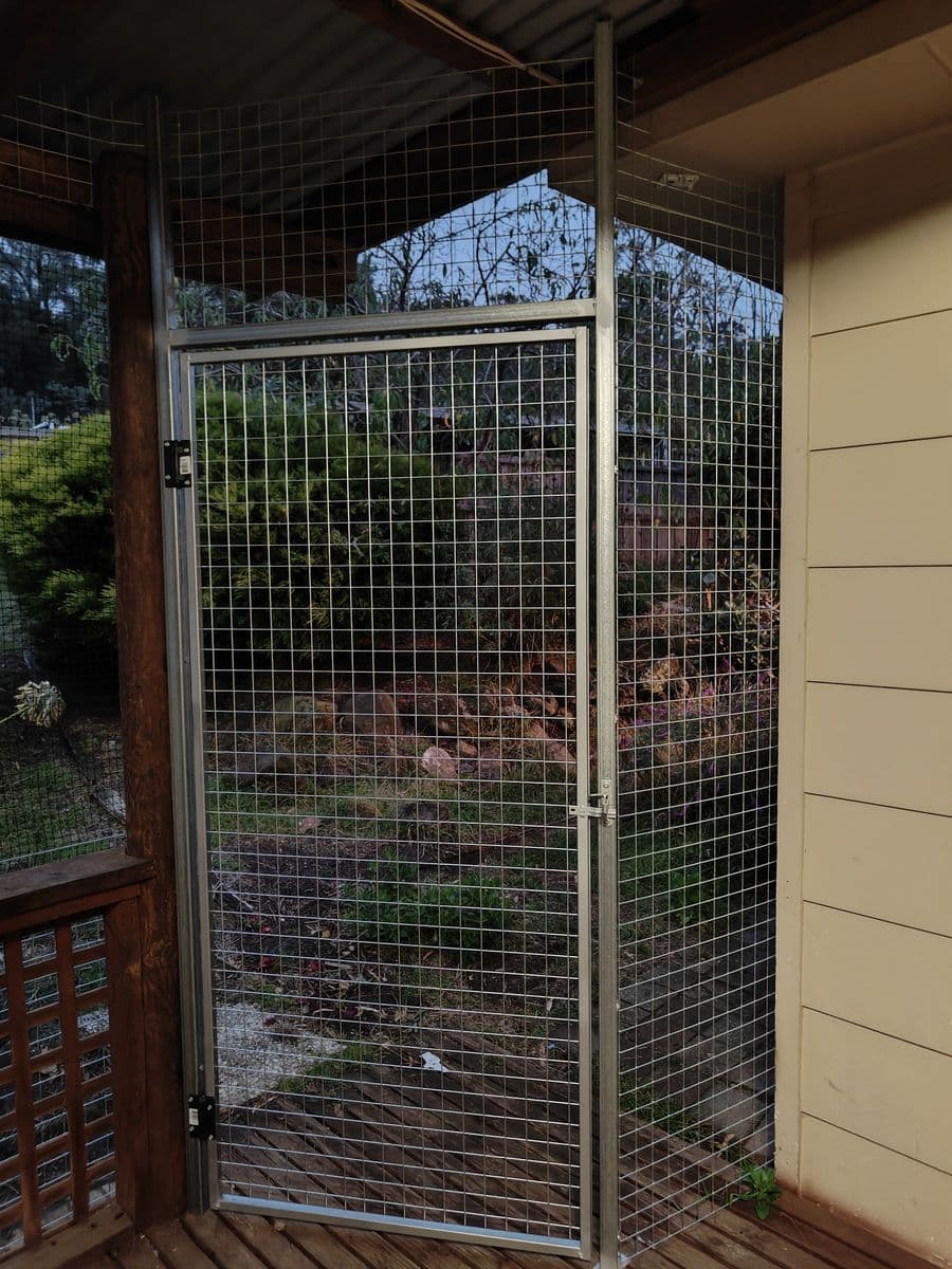 A galvanised mesh gate in a metal frame at the corner of a covered deck catio, looking out to the garden