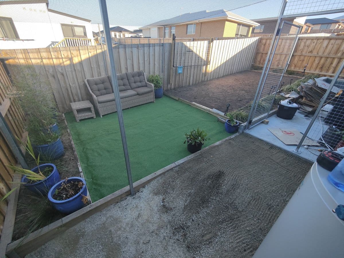 A netted back yard with a patch of artificial turf, an outdoor lounge and pot plants between paling fences