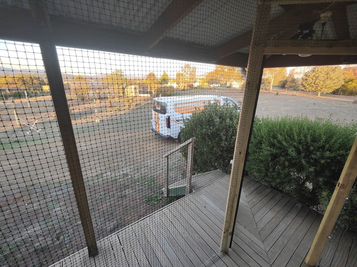 A timber verandah enclosed with black netting, looking out over shrubs to open ground