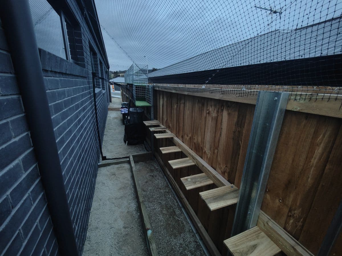 A long netted side run between a dark brick house and a timber fence, with a line of timber steps fixed to the fence
