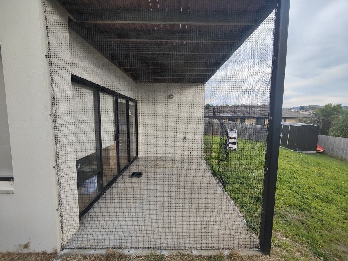 A concrete patio under an upper deck, enclosed with netting on black posts, opening onto a lawn