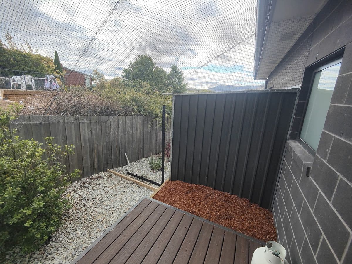 A small timber deck and mulched garden bed under netting beside a dark brick house and a paling fence