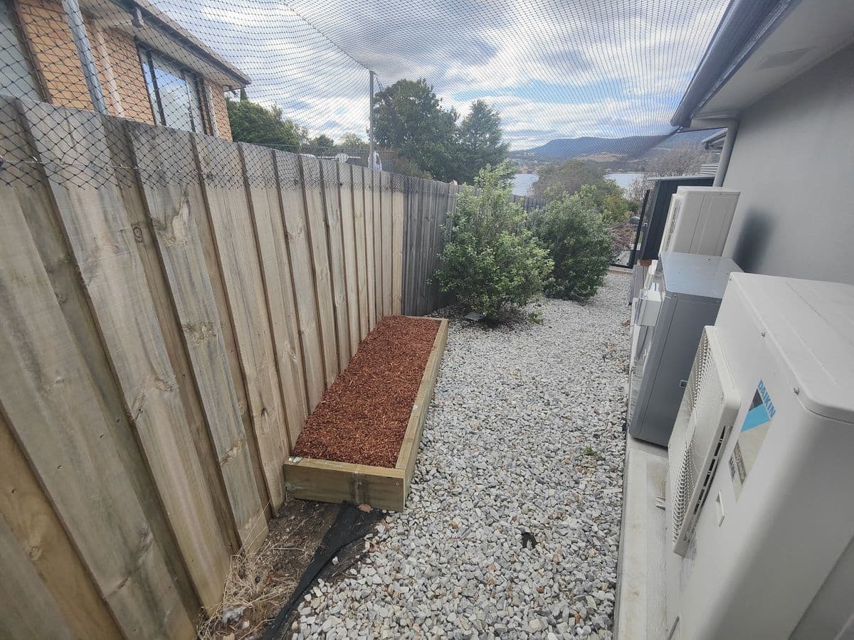 A netted side run with white gravel and a raised mulch bed between a timber fence and a house