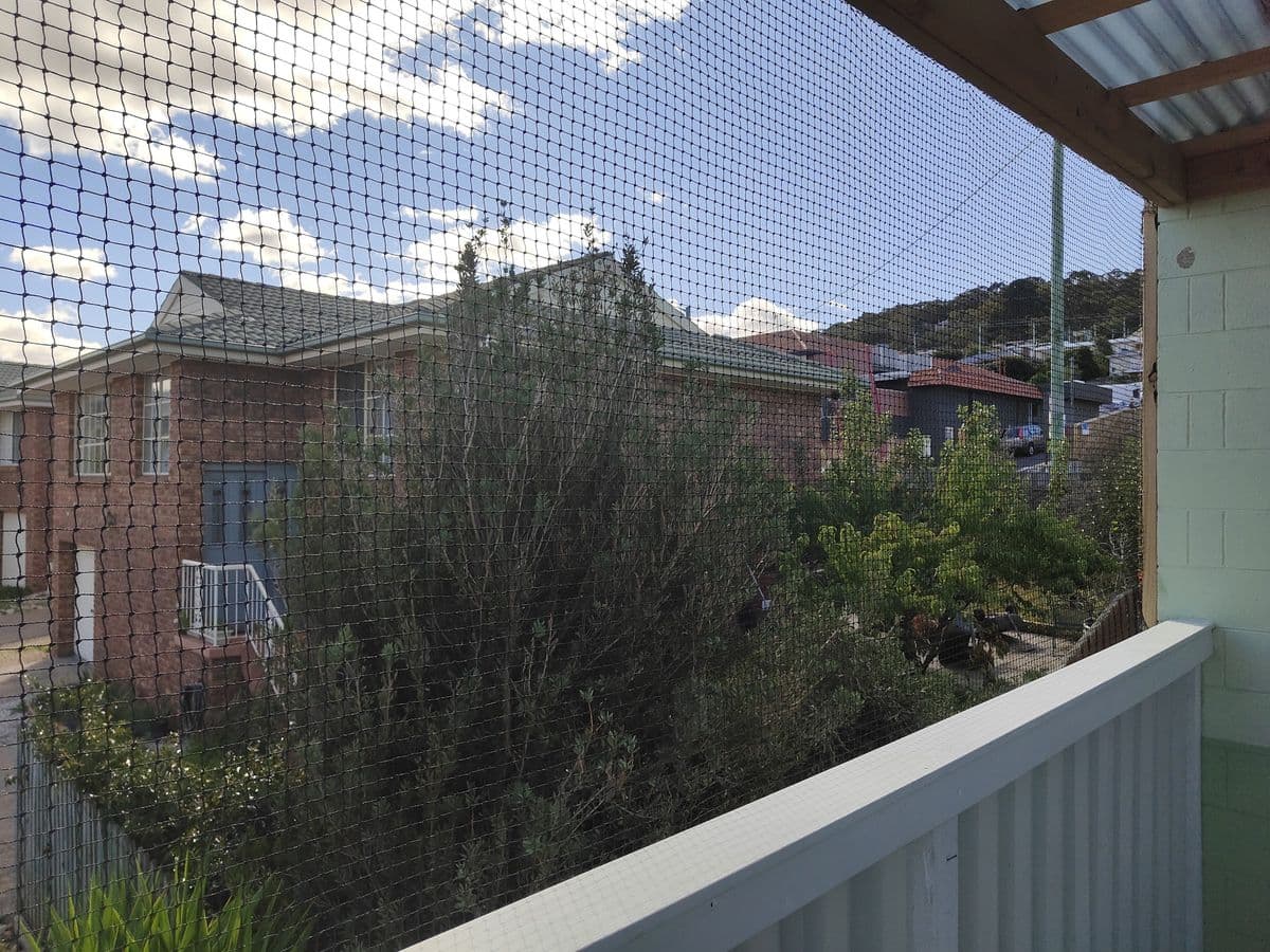 A balcony enclosed with black netting from rail to roof, looking out over shrubs and neighbouring houses