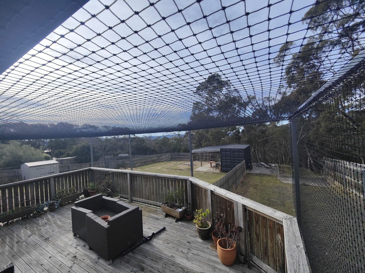 Black netting stretched over a timber deck with outdoor furniture and pot plants, looking out to bushland